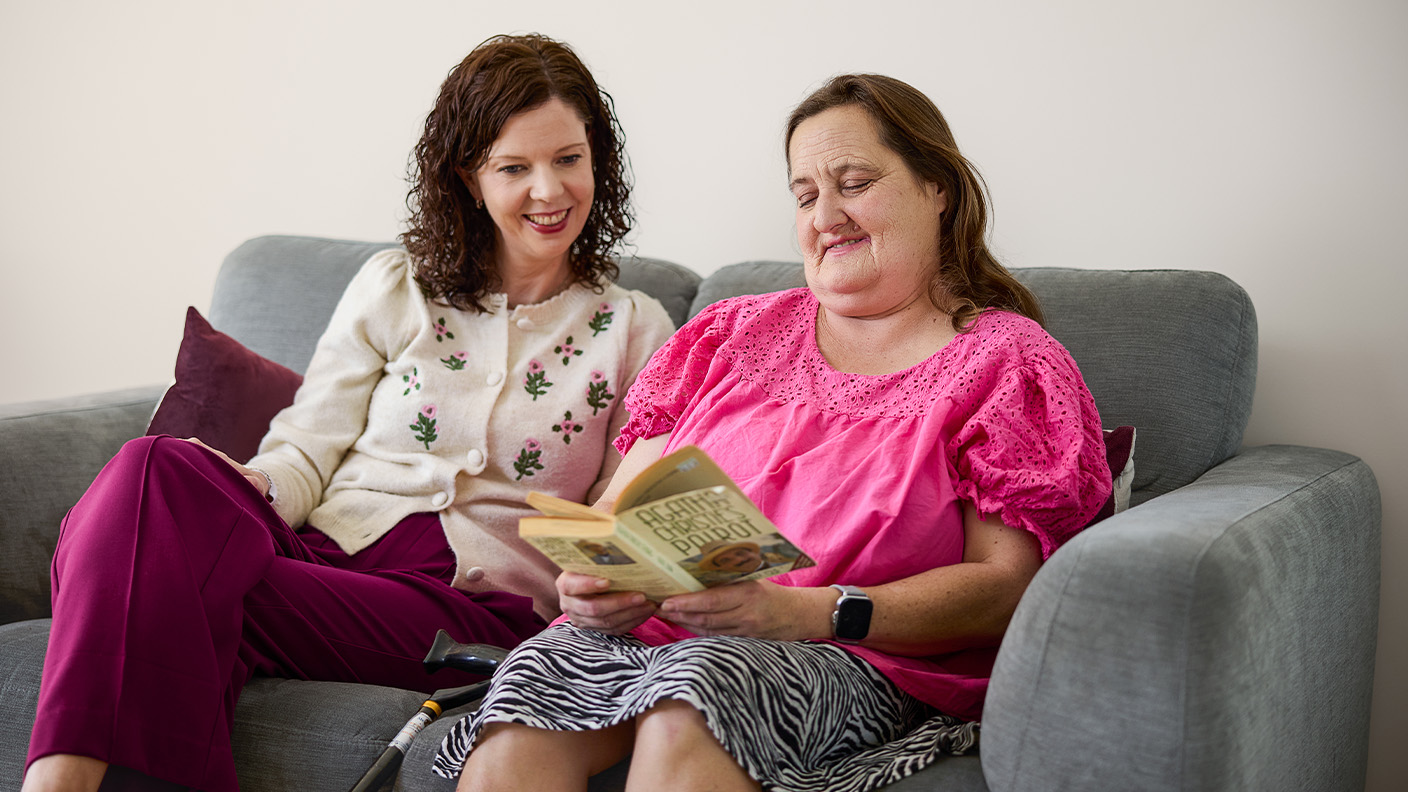 Two people are sitting on a lounge, looking at a book. One person has a disability.