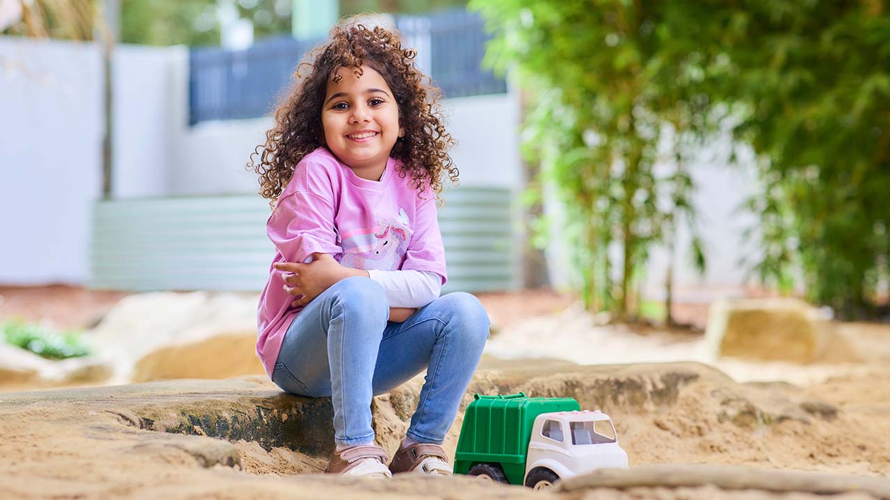 Child sitting in sandpit, playing with toy truck.