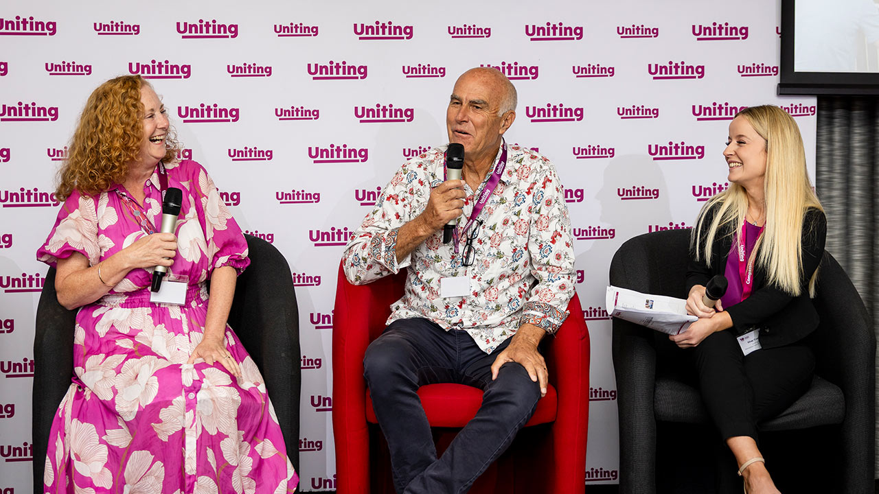 2 guests in conversation with Shay Costello from Uniting, sitting on stage and talking into hand held microphones.