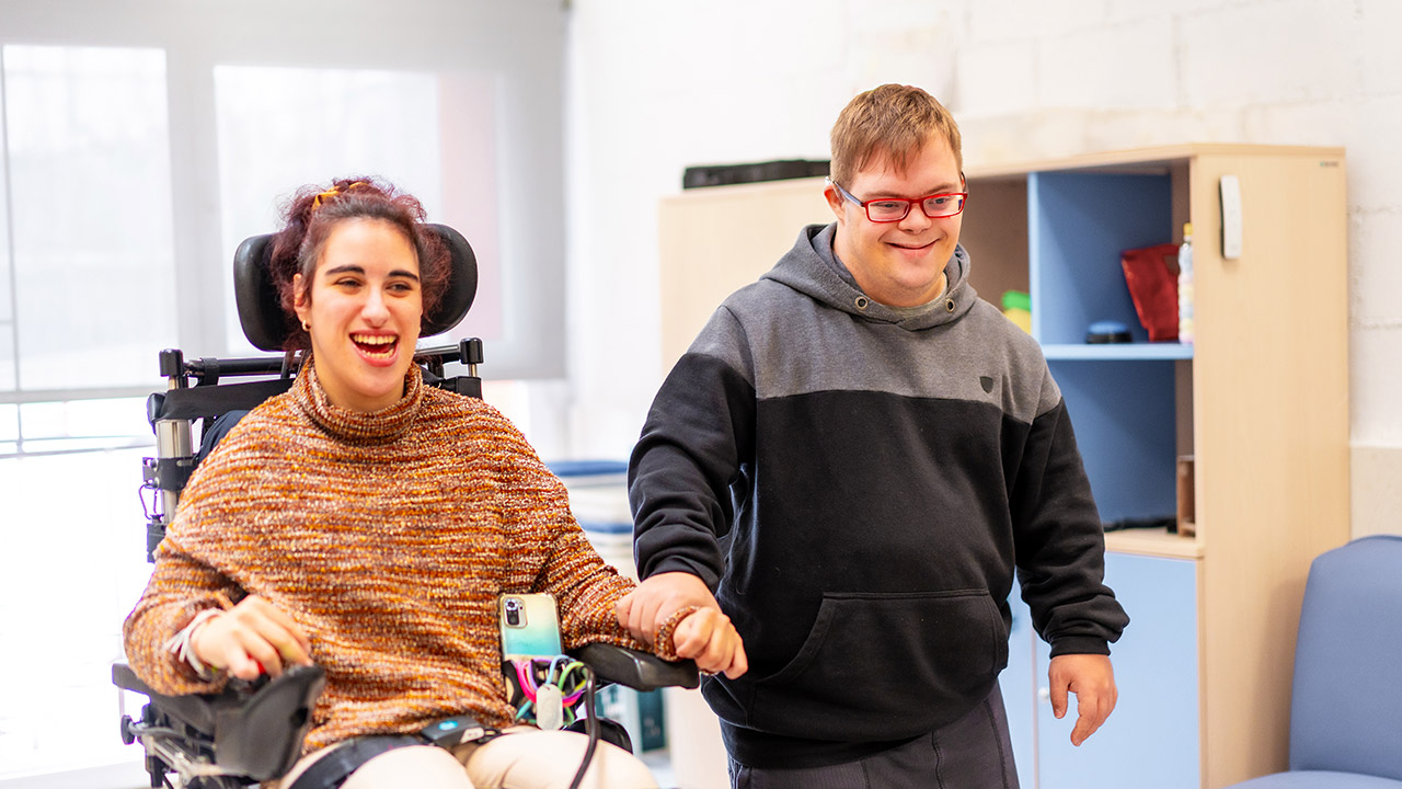 Two people with disability in a community centre. One person is sitting in a wheelchair, the other is walking beside them.