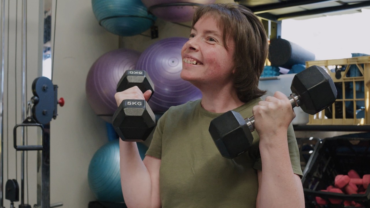 Angela is lifting weights at a gym and smiling.